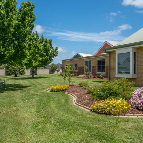 Well-maintained garden with colorful flower beds and green lawn outside single-story brick buildings under a bright blue sky with scattered clouds.