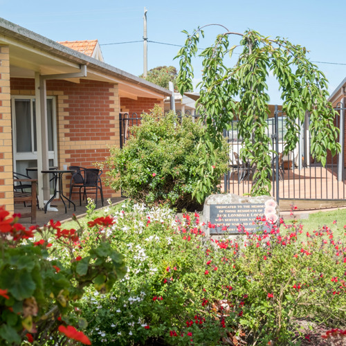 Jack Lonsdale Lodge Courtyard with red brick buildings, a garden of red and white flowers, a small drooping tree, and outdoor seating areas under covered patios, separated by a black metal fence. The scene conveys a peaceful, well-maintained communal space designed for relaxation and social interaction.