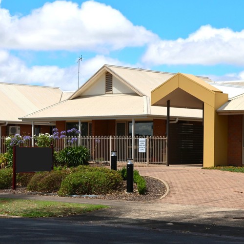 Single-story residential building with light-colored metal roofing, brick walls, and a yellow entrance feature. The property is surrounded by a low decorative fence and landscaped gardens with shrubs and flowering plants under a bright blue sky with scattered clouds.