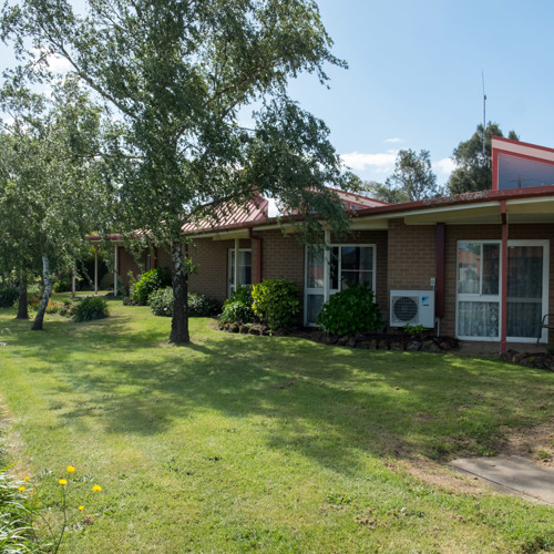 Single-story brick building with a red roof, large windows, and a covered porch, surrounded by green lawn, shrubs, and trees along a quiet street under a clear blue sky.