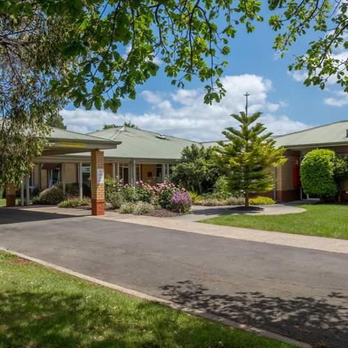 Talbot Place A well-maintained building with a green roof and brick walls is surrounded by lush greenery and colorful flower beds. The entrance features a covered walkway supported by brick columns. The scene is bright and sunny, with a clear blue sky and scattered clouds overhead. A paved driveway leads up to the entrance, flanked by manicured lawns and various trees, including a prominent conifer in the center of the garden area.