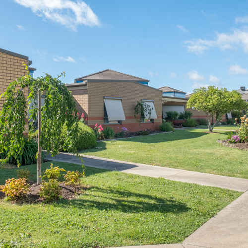 P.S. Hobson A suburban street lined with single-story brick houses, each with well-kept front lawns and gardens. The homes feature tan and red brick exteriors, some with window awnings. Small trees, shrubs, and flowering plants decorate the lawns. A concrete sidewalk runs along the street under a sunny sky with scattered clouds.