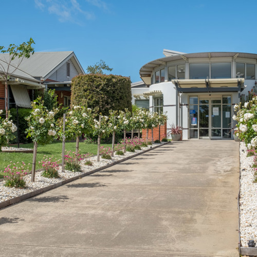 Modern building with circular entrance and large glass doors at the end of a paved walkway, lined with flowering trees and vibrant landscaping under a clear blue sky.